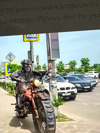 A weathered statue of a person in a hat and goggles rides a motorcycle on a city street. The statue is dark and appears to be made of metal. Behind the statue, several cars are parked along the road, and a pedestrian crossing sign is visible. The sky is blue, and trees line the background. The statue's attire suggests a vintage or steampunk aesthetic.
