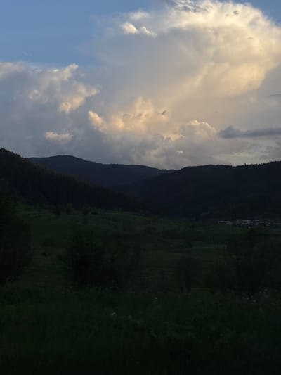The image shows a landscape in Borino, Bulgaria. On the left, there is a bright, cloudy sky, with shades of white, gray, and blue. In the middle, a dark mountain range stretches across the frame, covered in dense trees. On the right, there is a green landscape, possibly a field or meadow, partially obscured by shadows and trees. Some buildings can be seen near the top of the right-hand slope. The image is rotated 90 degrees.