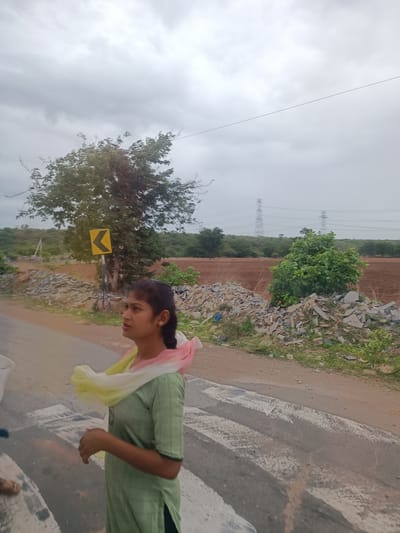 In Sugali Tanda, India, a young woman stands on a zebra crossing with a light green kurta and a multicolored scarf draped around her neck. She is facing to the left, with her hair pulled back in a braid. The environment is outdoors with a cloudy sky. Behind her, a large tree with dark green leaves stands near a yellow directional sign pointing left. The ground is a mix of dirt and rubble, with a brown field visible in the distance. Power lines and transmission towers are also present in the background, indicating a semi-rural or developing area. The road surface has prominent white painted stripes of the zebra crossing.