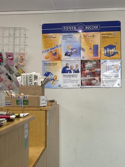 The image shows a wall in what appears to be a branch of the Russian postal service (Почта России). The wall has a blue sign with the name of the postal service in Russian and a number of promotional posters attached below. These posters advertise various services offered, such as Express Mail Service (EMS), payment of utility bills, vehicle insurance (ОСАГО), joining the postal service team, subscribing to newspapers and magazines, and receiving pension payments at home.

Below the posters, there is a wire rack filled with postcards and other small items for sale. Next to the wall is a wooden shelf or counter with various items stacked on it, including cardboard boxes, a stack of the newspaper "Gazeta Cherepovetskogo Rayona" (Газета Череповецкого Района), and other miscellaneous objects. The boxes appear to be from various manufacturers, including one with the brand "Jacobs" visible. The location is likely in the city of Тоншалово, Russia, given the newspaper publication area.
