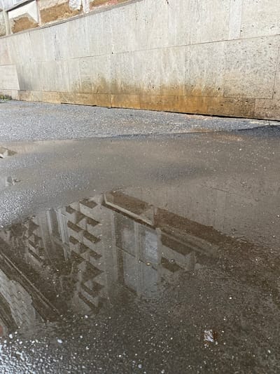 The image shows a puddle of water on asphalt next to a stone wall. The wall appears to be part of a building or a retaining wall. The asphalt is wet and reflective, showing a distorted reflection of a building with a patterned facade. The stone wall is made of large, rectangular blocks and has some staining or discoloration at the bottom. There is a strip of gravel between the asphalt and the wall. The scene is outdoors in daylight.