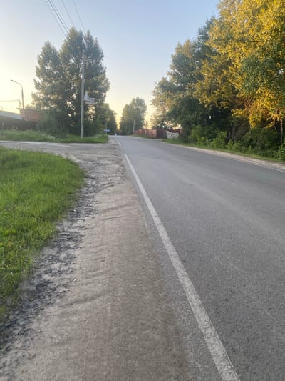The image shows a paved road in Semyonovskoye, Russia, at dusk. A grassy embankment borders the left side of the road, which is marked with a white line. Trees line the right side of the road, some of which are illuminated by the setting sun. A utility pole with a sign is visible on the left side of the road. The sky is clear and a pale orange, indicating the sun is low on the horizon.