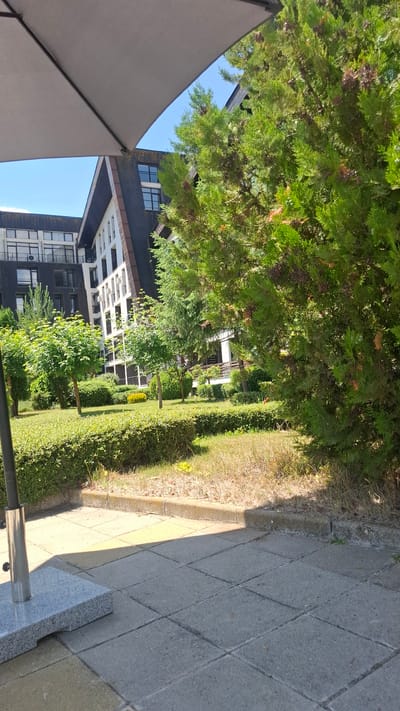 The image depicts a sunny day in the city of Razlog, Bulgaria, focusing on a courtyard area with a modern building in the background. A large, grey umbrella is partially visible in the foreground, casting a shadow on the paved patio made of square grey and yellow tiles. To the right of the patio, a well-maintained garden area is present, featuring trimmed green hedges and various trees, including evergreen conifers. In the background, a multi-story building with a distinct architectural style, characterized by a dark, angled roof and white facade with numerous windows, stands against a clear blue sky. The building appears to be residential or commercial, with balconies and air conditioning units visible. The overall scene suggests a tranquil, well-kept outdoor space, likely associated with the modern building.