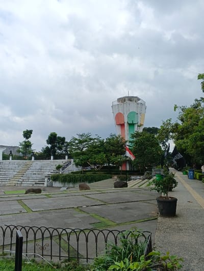 This image captures an outdoor public space in Bien Hoa, Vietnam, under an overcast sky. A tall, cylindrical tower dominates the right-center, featuring a hexagonal top and vertical panels painted in white, orange, light blue, and light green. To the left of the tower, a multi-tiered, white concrete amphitheater ascends a gentle slope. The foreground consists of a low, dark metal fence with arched sections, behind which lies a large, segmented paved area with concrete slabs separated by strips of grass. Several large, dark, smooth stones are scattered across this paved surface. A red and white flag is displayed on a pole near the base of the tower. Abundant green trees and vegetation are visible throughout the scene, notably surrounding the tower, behind the amphitheater, and lining a narrow, light-colored paved pathway on the far right. Two blue trash bins and multiple black potted plants are located along this pathway. In the upper left background, faint dark silhouettes of at least two statues are present on an elevated platform. A single small bird is visible in the upper right portion of the cloudy sky. No individuals are present in the depicted scene.