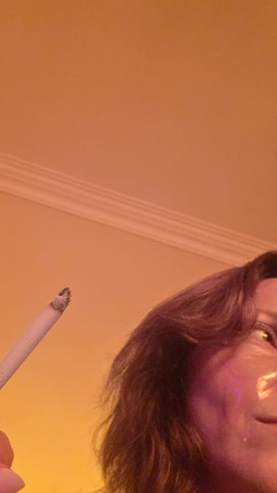 In Al Hadaba, Egypt, a close-up shot captures a person holding a lit cigarette in their left hand. The cigarette is positioned in the lower-left quadrant of the frame, with its burning end clearly visible. The person's face is partially visible on the right side of the image, with their hair obscuring some of their features. They appear to be wearing a face mask or cosmetic patches. The background is a plain, orange-toned wall with decorative molding near the top of the frame. The lighting is warm and diffused, creating a soft glow throughout the scene.