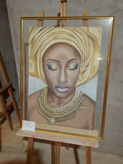 A framed painting of a woman wearing a yellow headscarf and a decorative necklace is displayed on an easel. The painting features the woman's face with closed eyes, highlighted by blue and yellow eyeshadow. A handwritten note is attached to the bottom left corner of the frame. The background appears to be a textured wall. The overall scene is set indoors, likely in an art gallery or exhibition space, given the presence of the easel and the presentation of the artwork. The painting itself is the central focus, with details of the subject's attire and facial features clearly visible.