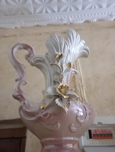 A decorative, ornate porcelain vase with a curved handle and floral embellishments sits in front of a beige wall. The vase is predominantly pink and white, with gold accents on the floral details. The top of the vase flares out with sculpted white fronds. Above the vase, there's an elaborately patterned white ceiling trim. A portion of a dark wooden cabinet and a white electrical panel are visible in the background.