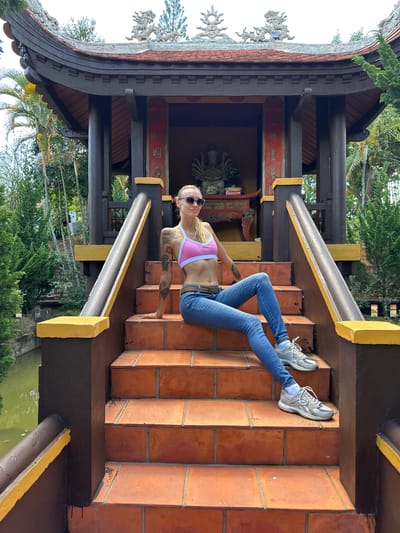 This media file features a young woman with blonde hair and sunglasses, wearing a pink sports bra and blue jeans, sitting on a set of red brick stairs. She has tattoos on her arms. The stairs are part of an outdoor structure with a dark brown and yellow color scheme, reminiscent of East Asian architecture, featuring decorative elements like dragon carvings. Behind her, a glimpse of a statue can be seen within an alcove adorned with red banners featuring Vietnamese text. The setting appears to be a temple or a culturally significant site in Vietnam, possibly Thôn 5. Lush green trees and foliage surround the structure, with a body of water visible in the lower portion of the image. The lighting suggests it is daytime, with sunlight casting shadows on the stairs, indicating fair weather. The woman's pose and expression convey a relaxed, casual demeanor.