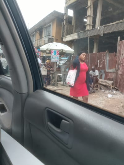 This media file captures a street scene in Alagbede, Nigeria, viewed from inside a car. The foreground is dominated by the interior of the car, with the door panel and window framing the outside view.

The primary subject in the mid-ground is a woman in a vibrant red dress, walking from left to right. She is carrying a white bag with a red logo and has a dark headscarf. To her left, a person in a yellow headscarf and patterned clothing is standing by a stall. The stall has a white umbrella and a sign for "POS SERVICES AVAILABLE HERE". The sign displays information about money transfer, deposit, and airtime services, with phone icons and text in English. Further to the right, a man in a light-colored t-shirt and brown shorts is seated on a wooden bench, seemingly resting.

Behind the subjects, the setting is a mix of unfinished construction and makeshift structures. An unfinished building with exposed concrete pillars and beams stands prominently, suggesting ongoing development. Nearby, corrugated iron sheets form a rustic fence. The ground is covered with loose gravel and dirt, with some scattered debris.

The lighting suggests it is daytime, possibly overcast given the diffused light. The overall atmosphere appears to be that of a busy urban or semi-urban environment, with commercial activity and ongoing construction.