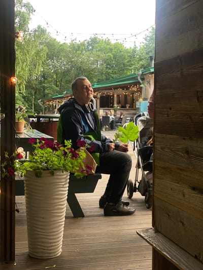 The image shows a man sitting on a bench outdoors. He is wearing a dark jacket over a green vest and dark pants. He has headphones around his neck and is holding a large green leaf. The man appears relaxed, looking to his right with his eyes partially closed. He is sitting in front of a planter with purple flowers. Behind him, there are more people, some sitting at tables, and a stroller. In the background, there are trees and lights strung between buildings. The scene suggests a casual outdoor setting, possibly a cafe or restaurant. The lighting and lush greenery indicate it is daytime and likely warm weather. The overall mood seems peaceful and unhurried.