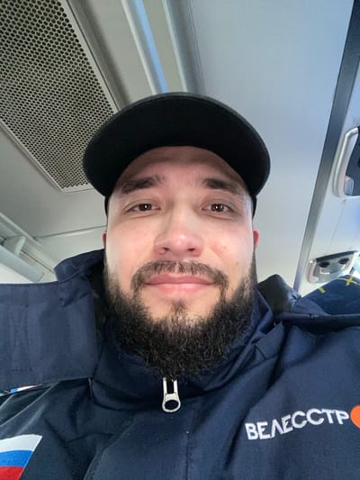 A selfie depicts a man with brown eyes and a black beard. He's wearing a dark blue jacket with the Russian flag and the Cyrillic word "ВЕЛЕCCTP" on it, and a black cap. The background includes a perforated panel and a window, suggesting he is inside a vehicle, possibly a bus. The city of Koskolovo, Russia is not identifiable in the image.