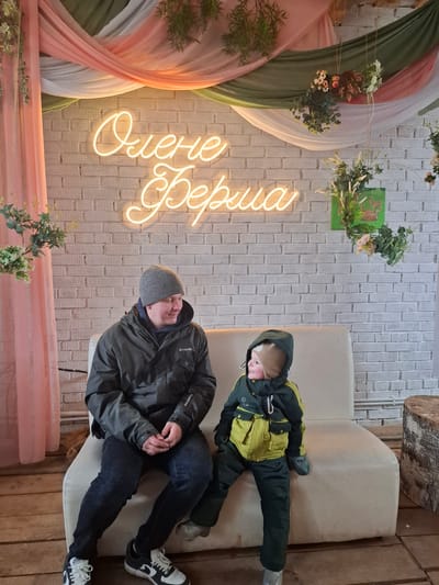 This image captures an indoor scene in Ludorvai, Russia, featuring an adult male and a young child seated on a light-colored, upholstered sofa or bench. The perspective is from slightly above and to the right, with the image rotated 90 degrees counter-clockwise.
The adult, in the foreground, is dressed in a dark winter jacket (a "Columbia" logo is visible on the sleeve), dark jeans, a grey knit beanie, and black and white sneakers. He is sitting with his hands clasped and appears to be looking downwards or to his left.
Behind him, the child is seated on the same sofa, wearing a hooded green and yellow winter suit and light-colored boots. The child is looking towards the left with a slight smile.
The background features a textured white brick-patterned wall. A prominent neon sign, glowing in a warm yellow hue, displays Cyrillic text that reads "Очень верила." This translates to "She very much believed" or "Very much believed."
To the left of the neon sign, decorative drapes in soft shades of pink, olive green, and white or cream are draped vertically. These drapes are adorned with artificial floral arrangements and green foliage, including a circular wreath-like decoration. Additional decor includes a small green panel with an animal silhouette (possibly a deer) and more foliage mounted on the wall. Rustic elements like a large wooden tree stump are visible in the upper right background, and wooden planks form the floor or a platform on the right side of the frame.
The warm clothing of both individuals suggests it may be cold outside, even though they are indoors. The scene appears to be a cozy or decorative waiting area, possibly in a cafe, event space, or a similar public establishment, blending rustic and modern decorative elements.