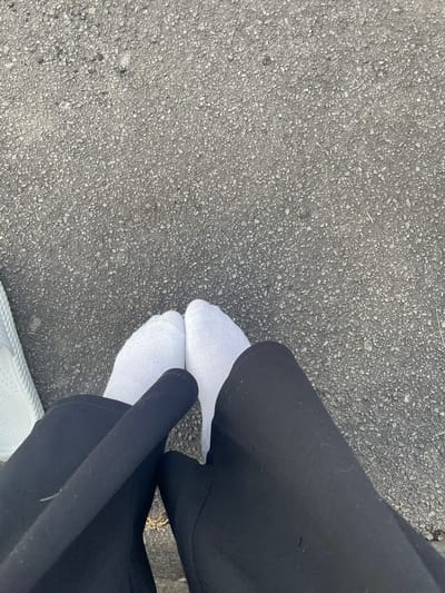 The image shows a close-up, overhead view of a person's legs and feet standing on asphalt. The person is wearing white socks and black pants. To the left of the frame, a portion of a white shoe or sandal is visible. The asphalt appears to be a typical road surface, with small stones and debris. There is no indication of the location in Wiehl, Germany within the image itself. The scene is static and focused on the lower extremities of an individual on an outdoor surface.
