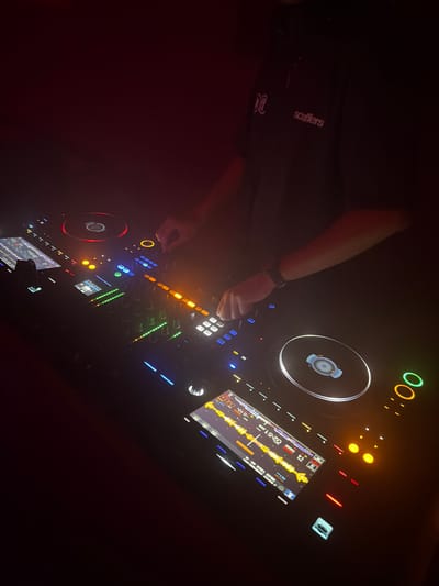 A person's arms are visible operating a DJ console in a dimly lit environment. The DJ console features multiple screens displaying waveforms and controls, with various illuminated buttons and knobs. The person is wearing a dark t-shirt with a logo and text on the left chest area. One hand is touching a control on the right side of the console, while the other hand is near a screen on the left. The background is dark with red ambient lighting. The scene is indoors, suggesting a club or performance venue. The image does not provide any information to identify the specific location within Hong Kong, China.