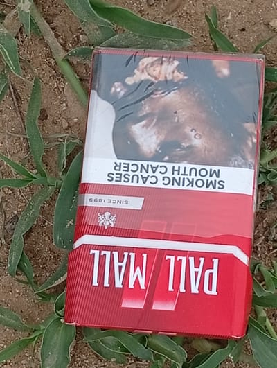 A red and white cigarette pack of Pall Mall brand cigarettes lies on the ground amidst dry dirt and green grass. The pack displays a graphic health warning image on its top portion, depicting a close-up of a person's face with visible lesions. Below the health warning, text reads "SMOKING CAUSES MOUTH CANCER." Further down, the text "SINCE 1899" is visible, along with a crest or logo. The bottom portion of the pack prominently features the brand name "PALL MALL" in large white letters. The scene appears to be outdoors in an unkempt area. The exact location in Gulak, Nigeria, cannot be determined from the image alone.