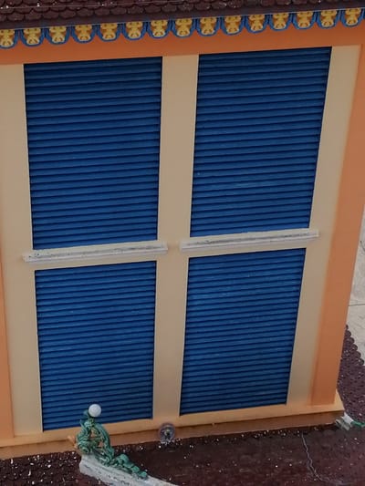 This is a close-up, slightly angled exterior shot of a decorative structure, likely part of a building or a miniature model, in Xuân Thành, Vietnam. The foreground is dominated by an orange-colored facade with two sets of blue louvered windows stacked vertically, separated by a horizontal white trim. The windows are uniformly blue and appear to be closed. The top of the structure features a dark brown, tiled roof with an orange band below it, adorned with a repeating pattern of yellow decorative elements. A small, green, stylized decorative object with a white sphere at its top is visible in the lower left corner, resting on a white base. The ground beneath the structure is a dark, textured surface. The overall impression is of a brightly colored, ornamental architectural detail.