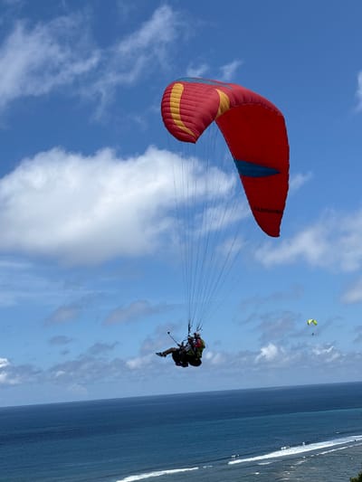 A person is paragliding over the water near the city of Benoa, Indonesia. The paraglider has a red, yellow, and blue canopy. The person is wearing a black harness and black shoes. The sky is blue with white clouds. The water is blue. There is another yellow paraglider in the distance. Some trees are seen to the right of the image.