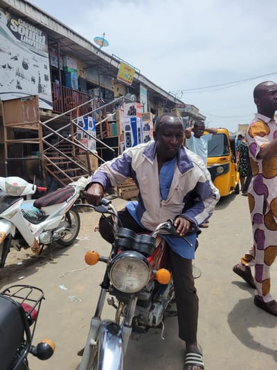 The image depicts a man sitting on a motorcycle in what appears to be a busy street in Suleja, Nigeria. The background features a building with electronics and musical instrument stores like "Calvary Sound Limited." In the foreground, a yellow tuk-tuk and several other motorcycles are visible. A man in a patterned outfit stands nearby. The overall scene suggests a bustling commercial area.