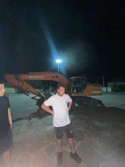 Two men are standing in a paved outdoor area at night. The man on the left is wearing a black t-shirt and blue shorts. The man on the right is wearing a white t-shirt and dark shorts. Behind them is an orange excavator with the brand name "DOOSAN" visible on its arm. The area appears to be a construction site or an industrial area, with a few buildings and light poles visible in the background. The lighting is artificial, likely from the light poles. The ground is paved with large, irregular stones. The overall scene is dimly lit, with the excavator and the men being the most prominent subjects.