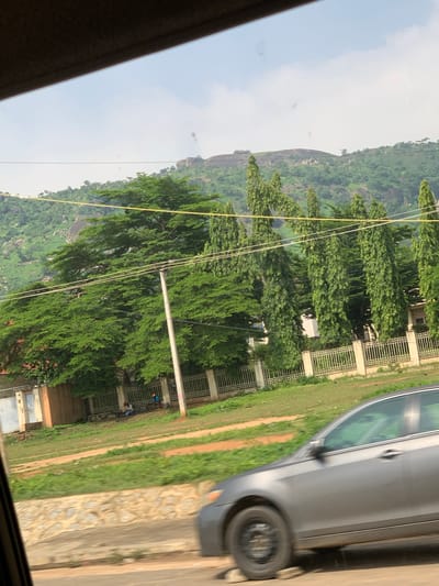 A vertical shot taken from inside a vehicle looking out at a lush, green landscape and a grey sedan parked on the right. The scene is likely Dam Camp, Nigeria. A steep, rocky hill covered in dense vegetation dominates the background. In the middle ground, a row of tall, mature trees with vibrant green leaves stands in front of a building with decorative metal fencing. Several utility poles and wires run across the scene, interspersed with the trees.
On the right side of the image, the grey sedan is partially visible, showing its side and rear wheel. The car's interior appears to have a green tint. Adjacent to the car and the building, there is a patch of grass and a dirt path. A few people, appearing as small figures, can be seen in the distance near the fenced building, suggesting some activity or presence. The overall impression is one of a natural, somewhat undeveloped setting with some human infrastructure present. The lighting suggests it might be daytime, possibly overcast given the diffused light. No visible text is present.