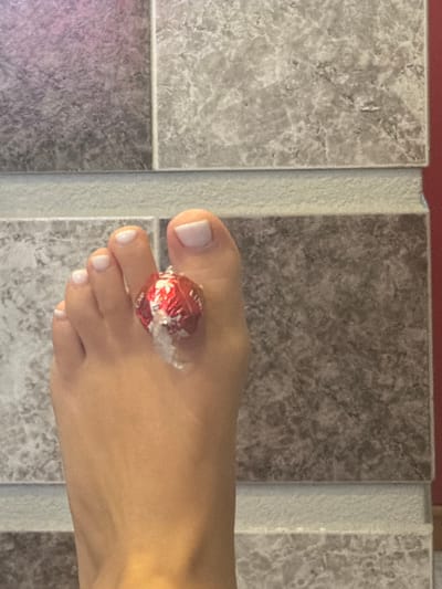 The image shows a close-up of a person's left foot, specifically the toes. A small, red, foil-wrapped candy with some white residue is positioned on the second toe from the big toe. The toenails appear to be clean and manicured with a pale polish. The foot is resting on a tiled surface. The background consists of tiled walls with a marble-like pattern in various shades of gray and a hint of pink in the upper left corner. The floor tiles are a lighter gray, and a thin strip of lighter textured material separates the wall tiles from the floor tiles. The scene is indoors, with the lighting suggesting it could be a bathroom or a similarly tiled area. There is no indication of the location in Novi Iskar, Bulgaria.