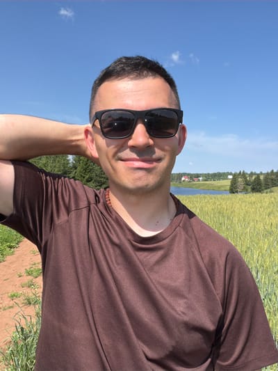 In Podshivalovo, Russia, a man is captured in a selfie-style portrait outdoors. He is wearing a brown t-shirt and black sunglasses. His right arm is bent, with his hand behind his head. The background features a clear blue sky with a few scattered clouds, a field of tall, green wheat, a body of water, and a cluster of houses and trees in the distance. The lighting suggests it is daytime, likely sunny.