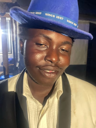 A close-up, eye-level shot shows a person wearing a blue fedora hat tilted to the left. The hat has a blue band with white text that reads "since 1887" repeated. The person has dark skin and is looking towards the camera with a slight smile, revealing their teeth and a hint of pink tongue. They are wearing a white collared shirt with vertical pleats on the placket, layered under a cream-colored vest or jacket with black lapels. The background is dimly lit and appears to be an indoor setting with vertical wooden beams and possibly some reflective surfaces on the left. The lighting is uneven, with a strong light source to the left of the frame creating highlights on the person's face and clothing. There are no visible signs of the location being Wumat, Nigeria.