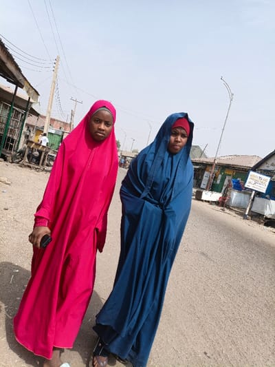 Two individuals are present on a paved street in Gawuna, Nigeria, under a clear daylight sky. The person on the left wears a hot pink full-body garment and a matching headscarf, holding a black smartphone in their right hand. Their face is visible, exhibiting a neutral expression. The person on the right wears a dark blue full-body garment over a red head covering. Their face is also visible, displaying a neutral expression. Both are positioned on the road surface. The background on the left includes several market-style structures with corrugated roofs, a wooden utility pole with power lines, and a parked motorcycle. Distant figures are partially visible. On the right, further structures are present, including one with a partially legible sign indicating "MAMAYA TEA AND PROVISION STORE," alongside a metal street light pole.