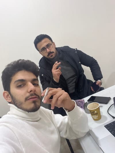 The image depicts two adult males indoors, located in Al-Mazza Municipality, Syria. Both individuals are seated at a white table with a plain, light-colored wall as the background.

In the foreground, a male with dark hair and a beard, wearing a white turtleneck sweater, holds a lit cigarette between the index and middle fingers of his right hand. He maintains direct eye contact with the camera. Positioned behind him and slightly to his right, a second male individual with dark hair, a mustache, and glasses wears a dark jacket over a grey sweater. He also holds a lit cigarette in his right hand and a dark-colored smartphone in his left, looking towards the camera.

On the white table surface in the lower right portion of the frame, several objects are visible: multiple Syrian Lira banknotes (including at least one 2000 SYP note), a dark-colored smartphone placed face down, a brown disposable coffee cup with text, a metallic ashtray containing ash, and the keyboard section of a silver laptop.