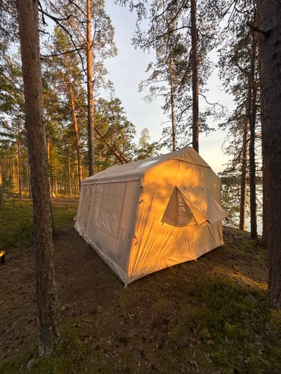 A large, tan canvas tent is situated in a forest clearing near a body of water. The tent is illuminated by the warm, golden light of the setting sun, casting long shadows. The surrounding environment consists of tall, slender pine trees with sparse green foliage and a forest floor covered in pine needles, moss, and some low-lying bushes. In the distance, a body of water, likely a lake or river, is visible, reflecting the soft light of the sky. The sky is a pale blue with hints of orange and pink, indicating the late hour of the day. There are no people or animals visible in the scene. The scene is peaceful and serene, evoking a sense of isolation and immersion in nature. The location is identified as Kutuzovskoe, Russia.