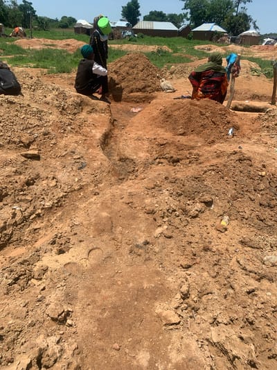In Buga da Gindi, Nigeria, a scene unfolds amidst dry, dusty terrain. Three individuals are present, engaged in activities that suggest manual labor or daily life. One person, dressed in a black vest and green headscarf, is seated on a mound of reddish-brown soil, facing left. Another person, wearing a black garment with a green head covering, stands near a bright green bucket. The third individual, adorned in a colorful patterned dress and green headscarf, is also seated, facing away from the camera.

The environment is characterized by uneven, disturbed ground, with piles of excavated earth. In the background, simple structures with thatched or corrugated metal roofs are visible, indicative of a rural or semi-rural settlement. The overall impression is one of an outdoor work area or a communal gathering space in a dry climate. Scattered debris, including plastic and metal containers, are also present, suggesting human activity.
