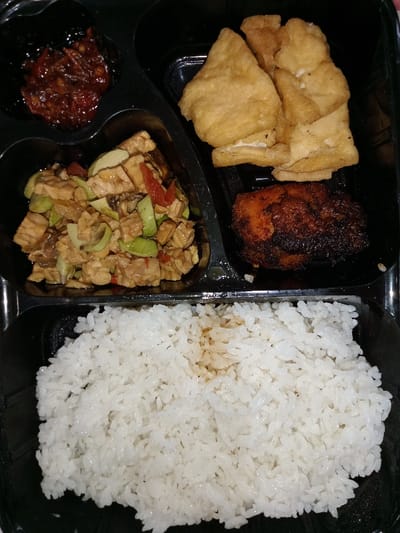 This media file displays an aerial view of a ready-to-eat meal presented in a black, partitioned takeaway container, indicative of Indonesian cuisine. The meal is divided into several compartments.

In the top-left compartment, there is a small serving of dark red sambal or chili paste, featuring visible chili flakes and possibly other spices, suggesting a spicy condiment.

The middle-left compartment holds a stir-fried dish, predominantly consisting of cubed tempeh and sliced green petai (stink beans). Red chili slices are also interspersed throughout the dish, which is coated in a dark, glossy sauce.

In the top-right section, several pieces of fried tofu are neatly stacked. The tofu has a light golden-brown exterior, indicating it has been deep-fried.

Below the fried tofu, in the bottom-right small compartment, a piece of chicken or meat, possibly grilled or fried, is visible. It has a rich, dark brown color with some charred edges, suggesting a flavorful and well-cooked preparation.

The largest compartment at the bottom of the container is filled with a generous portion of plain white rice. Some light brown liquid or sauce stains are visible on the surface of the rice, possibly from one of the other dishes.

The scene depicts a typical Indonesian meal packed for convenience, likely for lunch or dinner. The food appears freshly prepared, and the black container provides a stark contrast to the colorful dishes. No text is visible in the image. Given the location context of Situbondo, Indonesia, these dishes are characteristic of local fare.