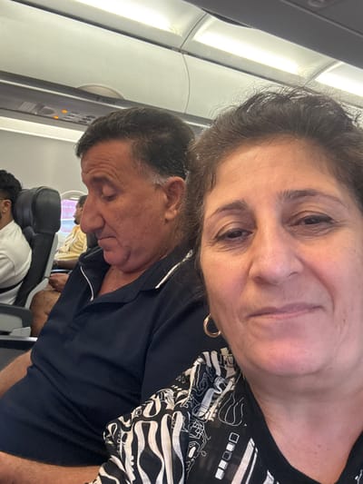 A medium shot captures a couple seated on an airplane, with the female in the foreground and the male to her left, slightly behind. The woman's face, positioned to the right of the frame, shows her looking towards the camera with a neutral expression. She has dark hair, brown eyes, and is wearing a black top with a white and black abstract pattern. A gold hoop earring is visible in her left ear. The man, to her left, is captured in profile, looking downwards. He has dark, graying hair and is wearing a dark blue polo shirt. Behind them, other passengers are visible in their seats, with airplane overhead compartments and lighting visible in the background. The overall scene depicts a moment during an airplane journey. The location in Sepang, Malaysia is not discernible from the image itself.