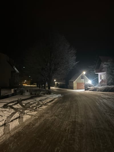 A night scene depicts a snow-covered road illuminated by streetlights. The road bends slightly, marked with tire tracks. Houses stand along the roadside, with snow-covered roofs and visible windows. Bare trees line the street, their branches stark against the dark sky. The image exudes a serene, wintry atmosphere.
