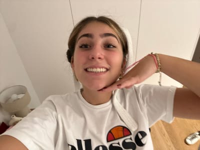 A young woman smiles directly at the camera. She wears a white t-shirt with the "ellesse" logo. Her hair is pulled back, possibly in a towel or headband. Her right hand is raised, with fingertips touching her chin in a casual pose. Bracelets adorn her right wrist. A bedside lamp is visible in the background, along with a glimpse of light-colored flooring. The backdrop consists of white walls.