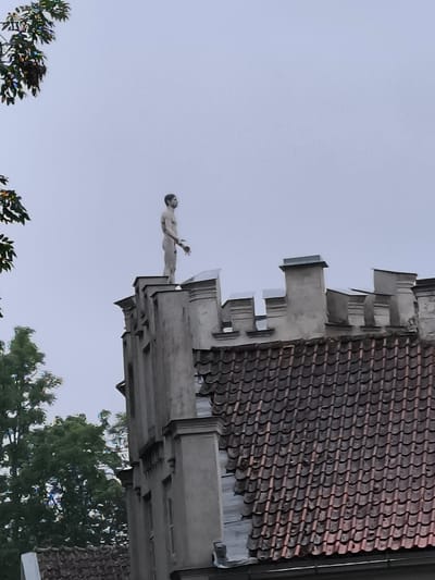 A full shot from below captures a pale, nude male statue standing at the apex of a section of a building's exterior. The statue, which appears to be made of a matte material, has dark hair and is depicted with its arms outstretched to its sides. The building itself is constructed of grey stone and features battlements along the top edge. The roof below is tiled in a reddish-brown color. To the left of the building, a lush green tree with dense foliage is visible, with some branches extending into the frame at the top. The sky is overcast and a pale blue-grey color. The scene appears to be located in Kuldīga, Latvia, based on the architectural style which is consistent with historic buildings in the region.