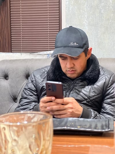 A man in a black jacket and cap holds a brown phone. He sits on a gray tufted chair. A wooden table with a shiny, dark object is beside him. A partially visible glass of amber liquid sits on the table. A wall with vertical brown slats and a textured gray wall are in the background. The image is slightly rotated.