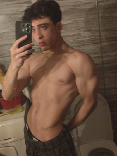 A young man takes a bathroom selfie, shirtless. He holds a smartphone in his left hand, obscuring part of his face. The man has dark, curly hair, and his gaze is directed at the phone. His body is toned, displaying defined abdominal muscles and biceps. The background reveals a typical bathroom setting: a tiled wall, a washing machine to the left with a red basket and yellow brush on top, and a partially visible toilet to the right. He wears patterned, low-waisted pajama pants.