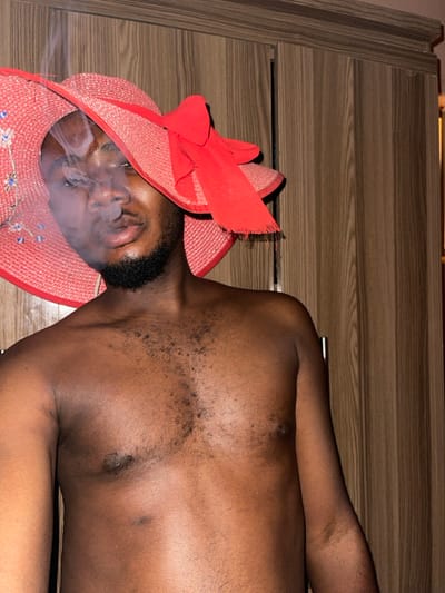 In Muchiya, Nigeria, an indoor portrait features an adult male. He is shirtless, displaying a hairy chest, and wears a red, wide-brimmed straw hat with a large red fabric bow and subtle floral embroidery. Smoke is visible emanating from the subject's mouth, partially obscuring his face as he looks towards the camera with pursed lips. The background consists of a light brown wooden wardrobe or cabinet with vertical paneling and a decorative top molding.