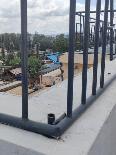 The image depicts an elevated view of a developed area within Juja ward, Kenya, under an overcast sky. In the foreground, a dark gray metal railing, composed of vertical bars welded to a horizontal base, is affixed to a weathered light gray concrete parapet or ledge, showing minor surface cracks.

The midground reveals a dense arrangement of structures. A multi-story building prominently features a significant vertical structural crack on its exposed light-colored exterior wall. Other buildings exhibit a mix of light blue and rusted corrugated metal roofs, interspersed with concrete and brick structures. Areas of dense green vegetation, including tall trees and banana plants, are scattered among the buildings. Utility poles and overhead lines are visible. Faintly, laundry is seen hanging on a line between two buildings.

The background consists of a distant tree line and subtle indications of hills or elevated terrain under a sky filled with white and gray clouds. No individuals, explicit actions, or direct interactions are visible.