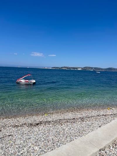 The media file depicts a bright, sunny day at Crveni Vrh, Croatia. The scene is a tranquil beach with clear, turquoise water lapping against a pebble shore. A white and red inflatable watercraft, possibly a towable tube or a small boat, is floating on the calm sea. In the background, a coastline dotted with buildings and lush green hills stretches along the horizon, suggesting a populated coastal town. The sky is a vibrant, cloudless blue, indicating fair weather and a warm day. The sun casts a bright light, illuminating the pebbles on the beach and the shallow areas of the water, where the seabed is visible. The overall impression is one of leisure and relaxation in a picturesque Mediterranean setting. There is no visible text in the media file.