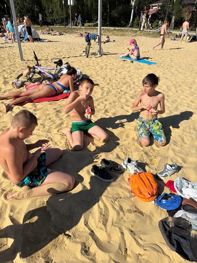 This image depicts a sunny day on a sandy beach, possibly in Malakhovka, Russia, given the provided context. In the foreground, three young boys are seated on the sand. The boy on the left is wearing patterned swim trunks and is looking down at his hands. The boy in the center is wearing green swim trunks with a "LACOSTE" logo and is eating from a small packet. The boy on the right is wearing colorful patterned swim trunks and is also eating from a packet. Scattered around the boys are several pairs of shoes, an orange backpack, and various folded clothes.
In the mid-ground, a woman in a blue bikini is lying on a red towel, facing away from the camera. A bicycle with a pink frame is parked nearby. Further back, other beachgoers are visible, some relaxing on the sand and others standing. The background shows a line of trees and some distant structures, suggesting a park or recreational area adjacent to the beach. The overall atmosphere is one of leisure and outdoor activity on a warm day.