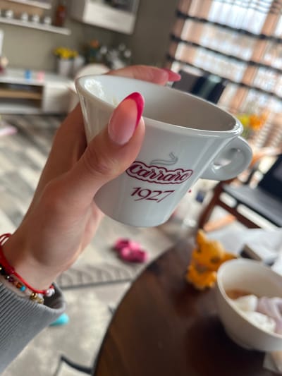 A person is holding a white Carraro 1927 coffee cup with red text and graphics. The cup appears to be empty. The person's left hand is visible, featuring a pink and clear french manicure. The person is wearing a gray long-sleeved shirt or sweater with a red beaded bracelet on their wrist. In the blurred background, there is a dark wooden table with a white bowl containing small white and purple items. Also in the background are windows with blinds. The location of Breznik, Bulgaria cannot be determined from the available image information.