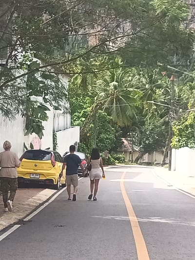 The image depicts a street scene in Ban Ao Nang, Thailand. A road with a yellow centerline curves into the distance, lined with white walls and lush tropical vegetation, including palm trees and other greenery. Three individuals are walking on the left side of the road, next to a yellow car parked on the shoulder. The foremost figure is an older person with gray hair, wearing a light-colored shirt and patterned pants. A man in a navy shirt and khaki shorts walks alongside a woman in a white outfit. A yellow car is parked nearby. Other vehicles are visible further down the road. The overall scene suggests a residential or tourist area with a relaxed atmosphere.
