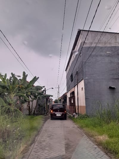 This image captures a narrow, paved street or alleyway in Kecamatan Gempol, Indonesia, under an overcast sky, suggesting a cloudy day or possibly recent rainfall, indicated by visible water droplets on the camera lens and damp-looking surfaces.
The street, paved with interlocking bricks or pavers, extends from the foreground into the distance. A dark-colored minivan or compact SUV, possibly maroon or dark brown, is visible in the middle distance, facing away from the viewer. Its taillights are illuminated, and a license plate is discernible on its rear, though the characters are illegible.
On the left side of the street, a large, two-story building with dark grey, somewhat textured walls dominates the view. Beyond it, other buildings of varying heights and lighter tones are visible, suggesting a residential area. Multiple electrical or utility wires stretch across the sky above these structures.
The right side of the street features a strip of green grass and scattered vegetation in the foreground. Further back, a cluster of large-leafed plants, appearing to be banana trees, grows alongside the road. A street light pole can be seen in the distance. No people are visible in the scene, and no explicit text is clearly legible besides the blurry license plate. The overall impression is that of a quiet, everyday urban or semi-urban environment.