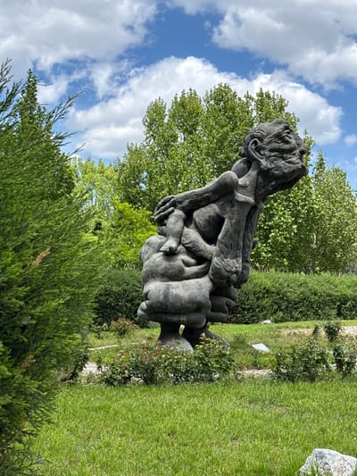 A sculpture is featured in the image, positioned outdoors in a grassy area. The sculpture, dark in color and seemingly made of stone or a similar material, appears to depict a human figure in a seated or contorted pose with its hands covering its face. The sculpture is situated amidst greenery, including bushes and trees. The background reveals a blue sky with scattered clouds. This scene is located in the Changping District, China.