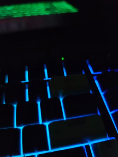 This is a close-up, dimly lit photograph of a mechanical keyboard. The keyboard features glowing blue backlighting for most of the keys, with some keycaps appearing a muted yellow or green. The top left of the image shows a blurred strip of green light, possibly from another screen or light source. A single, small green LED indicator is visible in the upper center of the frame. The overall impression is that of a gaming or computer setup in a dark environment. The image does not provide any information or context related to the city of Versailles, France.