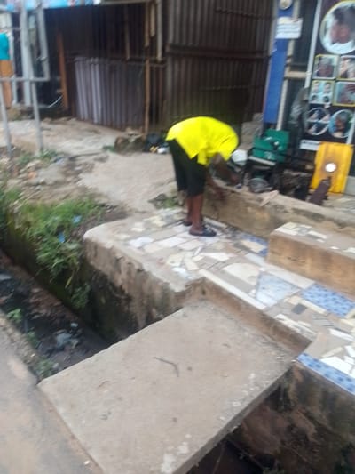 The image shows a person wearing a bright yellow shirt and dark shorts bent over a concrete structure. The person appears to be working or tending to something on the structure. The background reveals a setting that resembles a roadside shop or a service area, with corrugated metal structures and what looks like a display board with pictures. The ground is paved with tiles in some areas and dirt in others, with a visible drainage ditch on the left side of the image. There are some green plastic containers and a yellow object in the background, possibly part of the shop's inventory or equipment. The lighting suggests it might be daytime, but the sky is not visible, so the weather cannot be determined. The overall impression is that of a busy, possibly commercial, outdoor environment in a tropical or subtropical region, given the greenery and the type of structures. The image's focus is on the person and their activity, with the surroundings providing context.