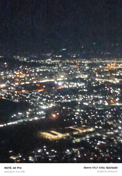 The image is a photograph of an aerial view of a city at night, with countless lights creating a bokeh effect. The lights are predominantly white and yellow, with some hints of orange and red, suggesting streetlights, building lights, and possibly vehicle lights. The overall impression is of a sprawling urban landscape seen from a distance, possibly from a high vantage point or from an airplane.
The bottom left corner of the image displays metadata: "NOTE 40 Pro" and "2026.06.21 11:44 PM". The bottom right corner shows camera settings and coordinates: "70mm f/1.7 1/13s ISO25600" and "40°28'2''N 49°56'41''E".
The coordinates provided, "40°28'2''N 49°56'41''E", point to a location in Azerbaijan, not Dushanbe, Tajikistan. This specific coordinate falls within the area of the Caspian Sea, east of Baku. Therefore, the image is not of Dushanbe. The scene depicted is a generalized urban night landscape. There are no discernible people, specific subjects, or objects that would indicate a particular activity or interaction. The environment is dark, with the city lights being the primary source of illumination.
