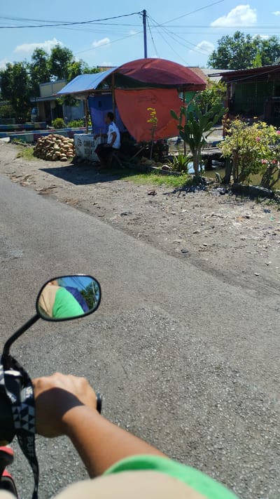 The image depicts a scene on a roadside in Cangkringsari, Indonesia. A person is seated on a wooden stool under a makeshift canopy covered with red and orange tarps, possibly a roadside stall. In front of the stall, a large pile of coconuts is stacked. The stall is situated next to a body of water, with a small wooden bridge visible. Several plants, including a cactus, are growing around the stall. The ground is a mixture of asphalt and gravel. The perspective suggests the photo was taken from a moving vehicle, likely a motorcycle, with the rider's hand and arm visible in the foreground. A motorcycle mirror reflects the rider and the sky. The background shows trees and buildings. The sky is clear and blue with some clouds.